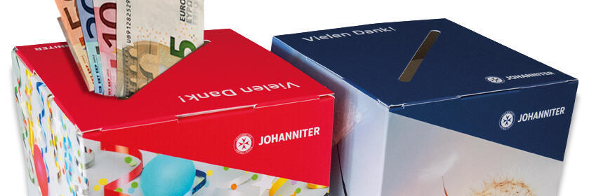 Zwei Johanniter-Spendenboxen, eine mit eingesteckten Euro-Scheinen, beide mit Aufdruck „Vielen Dank!“ und „Aus Liebe zum Leben“.