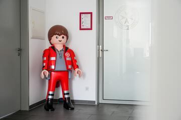 Kunststofffigur in roter Rettungsdienst‑Uniform neben Glastür mit Johanniter‑Logo.