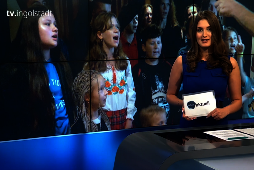 Nachrichtensprecherin im Studio von tv.ingolstadt präsentiert eine Sendung, im Hintergrund ist ein Kinderchor auf dem Bildschirm zu sehen.