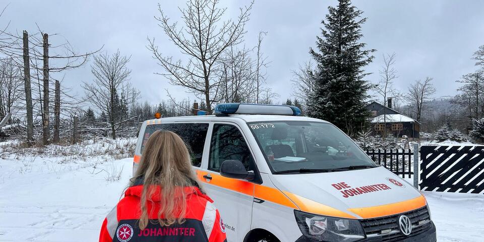 Eine Johanniterin von hinten vor einem Johanniter-Mannschaftstransportwagen in verschneiter Landschaft.