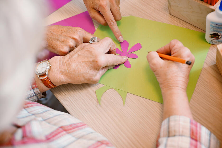 Eine ältere Person bastelt etwas auf einem Tisch. Eine weitere Person unterstützt sie dabei.