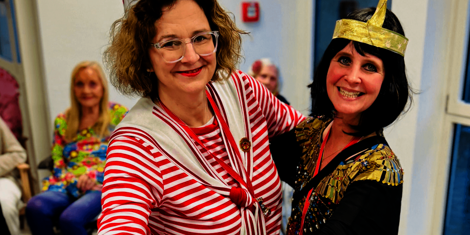 Zwei Frauen umarmen sich lachend; eine trägt schwarz-goldene Verkleidung, die andere gestreift.