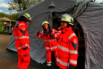 Drei ehrenamtliche junge Helfenden während einer Übung stehen vor einem Zelt und Behandlungsplatz für Verletzte. Alle drei tragen Einsatzkleidung in Orange und Sicherheitshelme. Eine Helferin zeigt beide Daumen hoch.