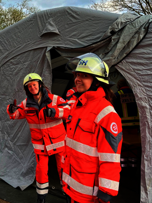 Drei ehrenamtliche junge Helfenden während einer Übung stehen vor einem Zelt und Behandlungsplatz für Verletzte. Alle drei tragen Einsatzkleidung in Orange und Sicherheitshelme. Eine Helferin zeigt beide Daumen hoch.