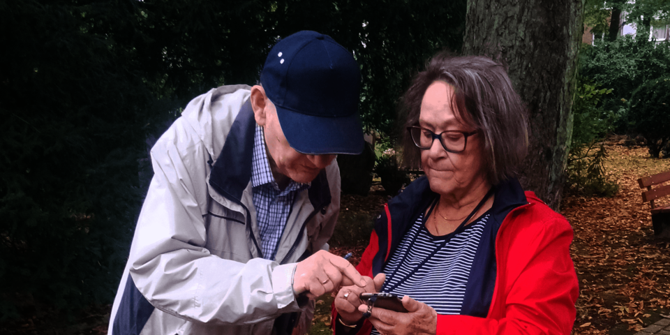 Eine Frau in einer roten Jacke steht im Freien und hält ein Handy in der Hand. Ein Mann neben ihr beugt sich darüber. Er trägt eine dunkelblaue Kappe.