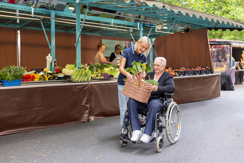 Eine Frau reicht einer älteren Frau, die in einem Rollstuhl sitzt, einen Korb mit Gemüse. Sie befinden sich auf einem Wochenmarkt.