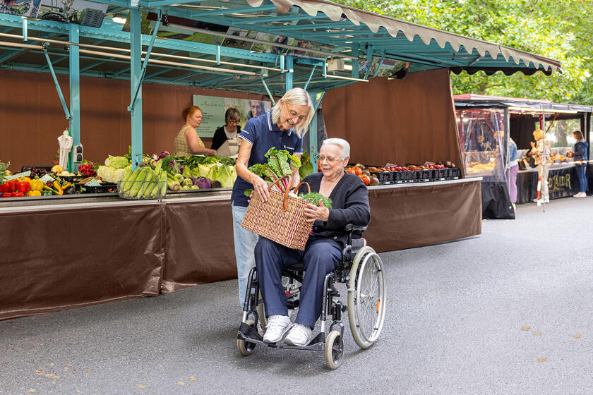 Eine Frau reicht einer älteren Frau, die in einem Rollstuhl sitzt, einen Korb mit Gemüse. Sie befinden sich auf einem Wochenmarkt.