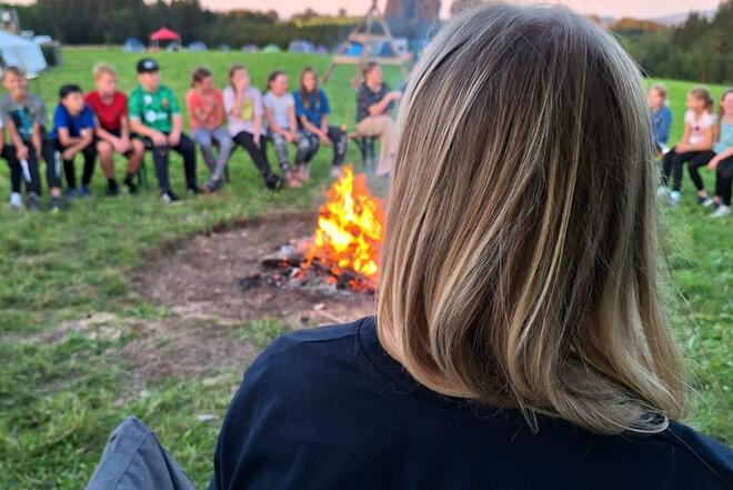 Man sieht eine Frau von hinten, auf ihrem T-Shirt steht „Zeltlagerteam“. Sie sitzt zusammen mit mehreren Kindern in einem Kreis um ein Lagerfeuer.