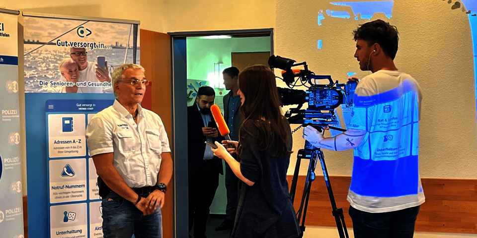 Ein Mann in Jeans und weuißem Hemd wird von einer Frau interviewt. Sie hält ein Mikrofon. Neben ihr steht ein junger Mann hinter einer Kamera