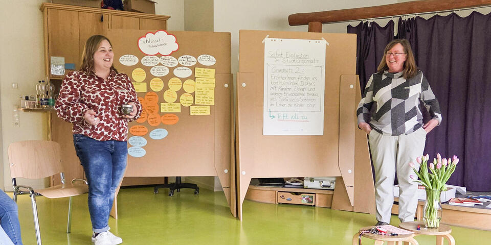 Zwei lächelnde Frauen vor zwei großen Pinnwänden auf denen ein Plakat und Zettel kleben. 