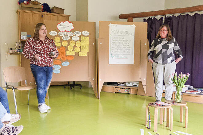 Zwei lächelnde Frauen vor zwei großen Pinnwänden auf denen ein Plakat und Zettel kleben. 