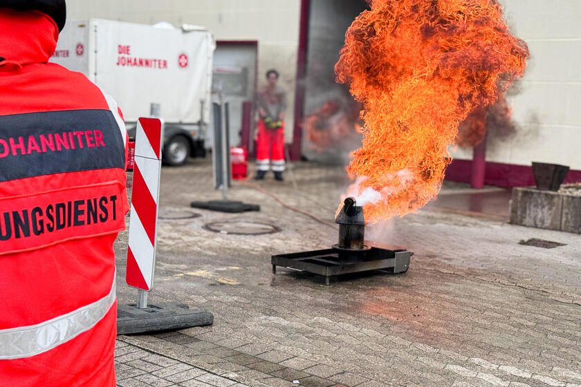 Eine Person mit Johanniter-Einsatzjacke ist im Vordergrund mit ihrem Rücken zu sehen. Im Hintergrund auf einem Parkplatz brennt ein Brandsimulator mit hoher Flamme. 
