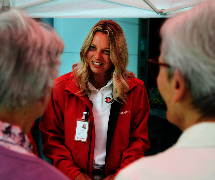 Eine Johanniter-Botschafterin in typischen Johanniter-Farben Weiß und Rot berät zwei ältere Personen.