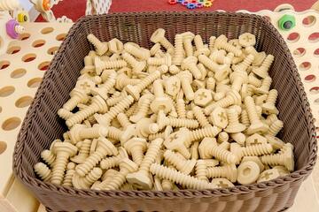 Brauner Korb mit großen Spielzeugschrauben. Im Hintergrund sieht man Holzgestelle in welche man die Schrauben schrauben kann.