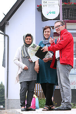 Ein Mitarbeiter der Flüchtlingshilfe an einer Bushaltestelle zeigt zwei Frauen mit Kopftuch den Weg anhand einer Karte.