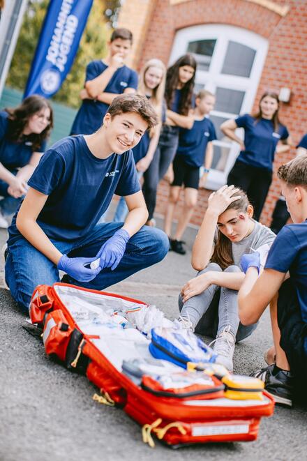 Jugendliche sitzen auf dem Boden vor einem Haus und üben Erste Hilfe zu leisten