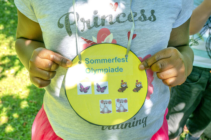 Ein Kind hält ein gelbes, selbst gestaltetes Schild mit verschiedenen Tieren abgebildet und der Aufschrift „Sommerfest Olympiade“.