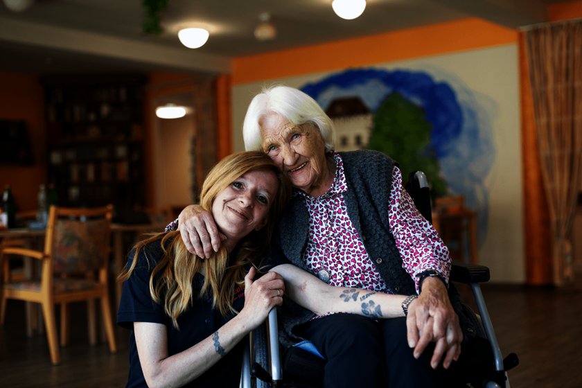 Pflegerin umarmt eine Seniorin im Rollstuhl, beide lächeln herzlich.  