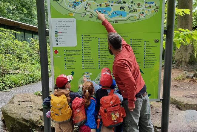 Mehrere Kinder stehen gemeinsam mit einem Erzieher vor einer großen Übersichtskarte im Tiergarten Nürnberg. Die Kinder tragen bunte Rucksäcke und schauen aufmerksam auf den Plan, während die Betreuungskraft auf einen bestimmten Punkt auf der Karte zeigt. 