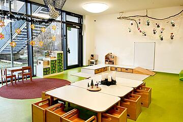 Heller Gruppenraum mit niedrigen Tischen, kleinen Stühlen, Spielmaterialien und großen Fenstern.