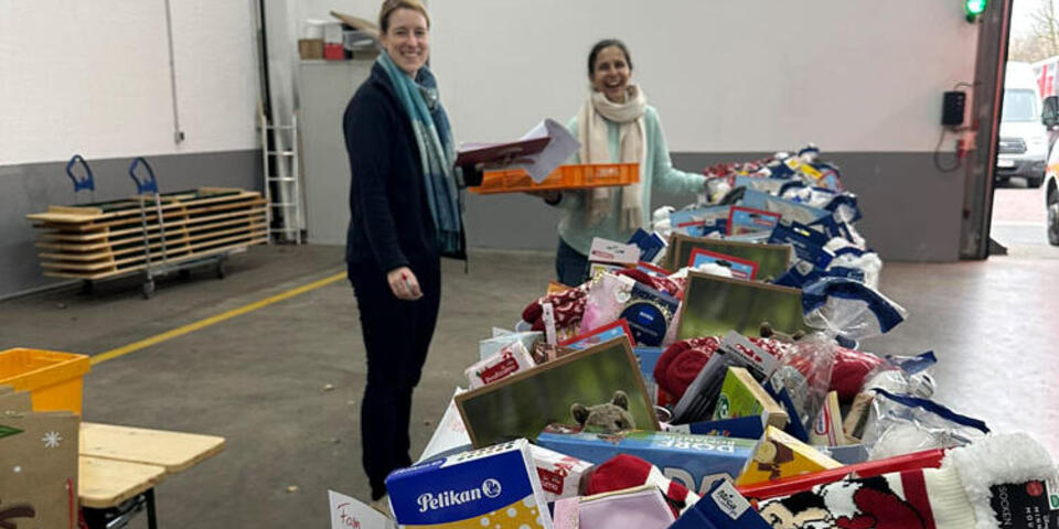 Körbe mit Spiel- und Schulsachen, Pflegeartikeln und Geschenken reihen sich hintereinander. Zwei Johanniterinnen stehen im Hintergrund und lächeln beim packen der Körbe für die Johanniter-Suppenküche. 