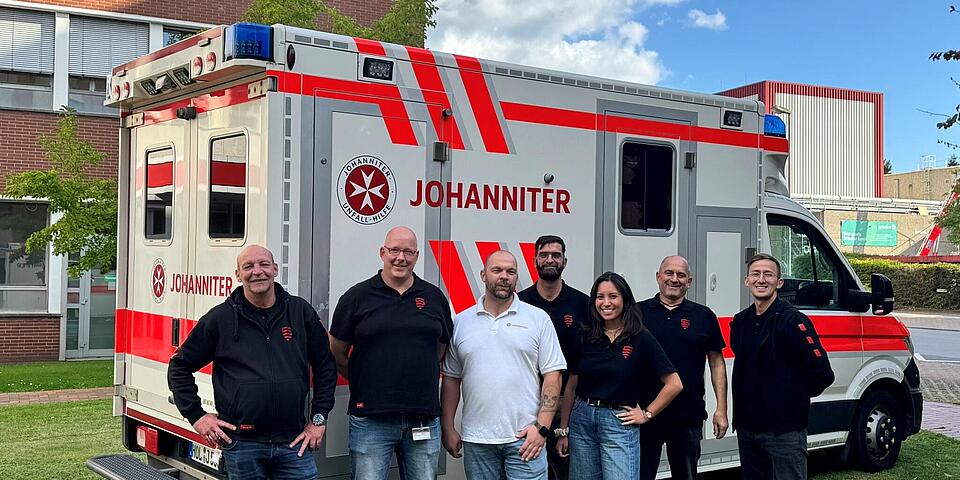 Fünf Männer und eine Frau mit schwarzer Oberbekleidung und ein Mann mit weißem Johanniter-Polo vor einem RTW der Johanniter