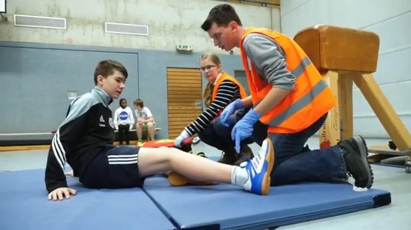 Ein Junge sitzt auf einer Sportmatte und wird von Schulsanitäter*innen versorgt.