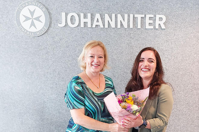 Zwei Frauen stehen vor einem grauen Hintergrund mit einem Johanniter Schild über ihnen. Die eine Frau überreicht der anderen einen Blumenstrauß.