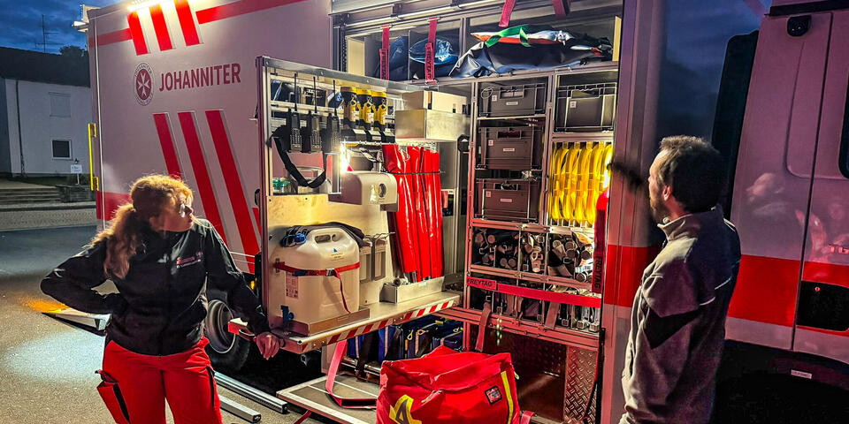 Ein Mann und eine Frau in Einsatzkleidung vor einem großen Gerätewagen. Die Seitentür des Fahrzeugs ist offen und man sieht viele Gerätschaften