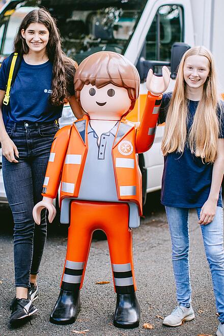 Zwei Jugendliche aus der Johanniter-Jugend stehen mit Playmobil-Johanniter vor einem Rettungswagen
