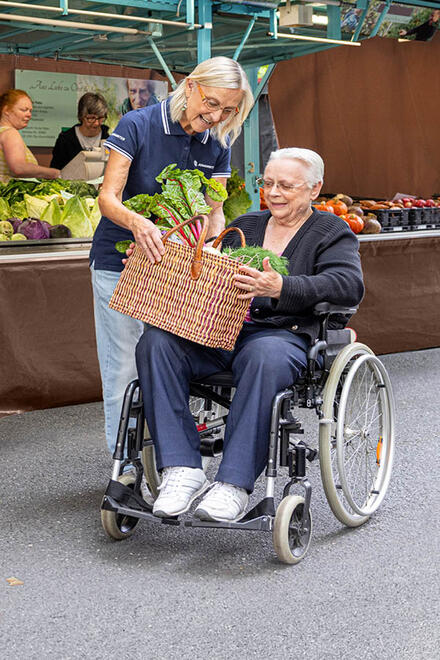 Eine ehrenamtlich Helfende versorgt eine Frau im Rollstuhl mit Gemüse auf einem Markt