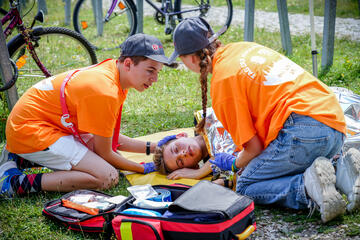Zwei Ersthelfer in Orange versorgen bewusstlose Person mit Rettungsdecke im Gras.