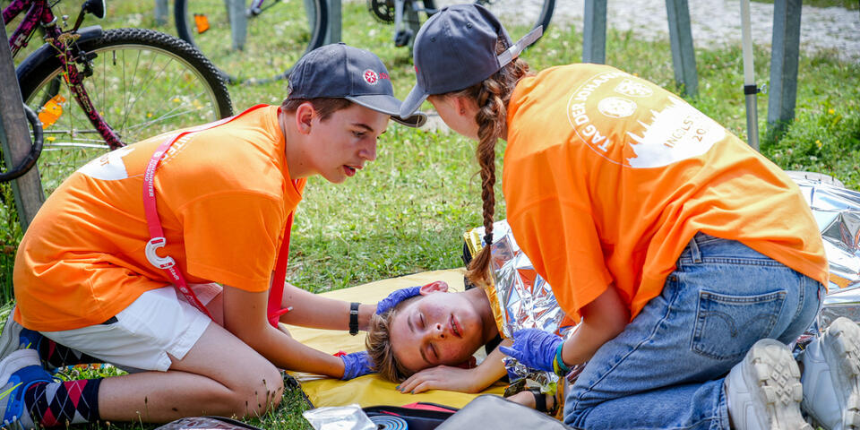 Zwei Ersthelfer in Orange versorgen bewusstlose Person mit Rettungsdecke im Gras.