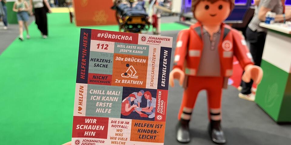 Eine Hand hält eine Postkarte. Im Hintergrund ist ein riesiges Playmobilmännchen in Johanniter-Kleidung zu sehen.