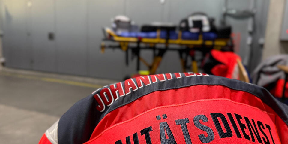 Eine Einsatzjacke mit Rückenschild auf dem steht "Sanitätsdienst" über einem Stuhl in einem Raum