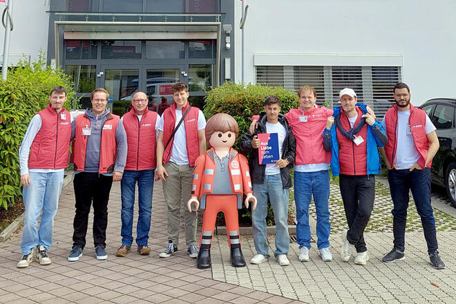Mehrere junge Leute in Johanniter Kleidung stehen vor dem Eingang zu einem Gebäude. Zwischen ihnen steht eine große Johanniter Playmobilfigur.