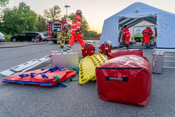 Medizinische Ausrüstung, Tragen und Notfallrucksäcke liegen vor einem aufblasbaren Zelt. Im Hintergrund bereiten Einsatzkräfte den Einsatz vor.