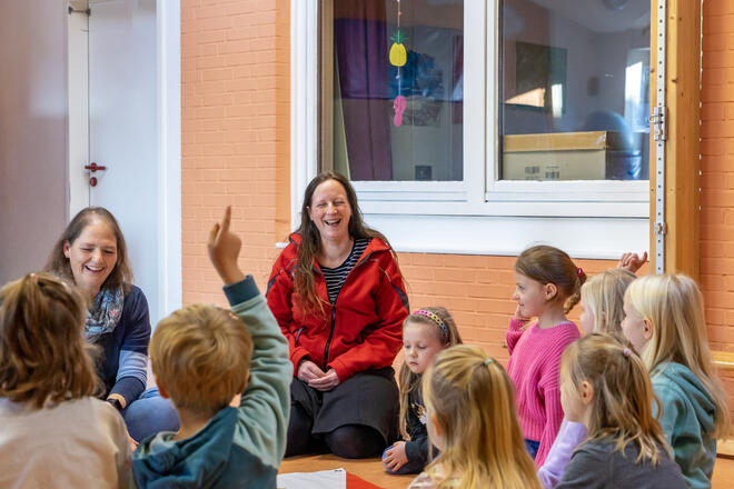 Zwei Erzieherinnen der Johanniter und mehrere Kinder sitzen in einem Kreis zusammen, ein Kind hebt die Hand.