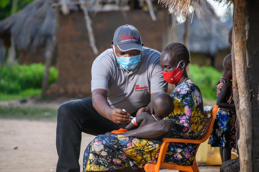 Ein Mann mit Johannitershirt misst den Oberarm eines Kindes das bei seiner Mama auf dem Schoß sitzt. Alle tragen einen Mund-Nasen-Schutz