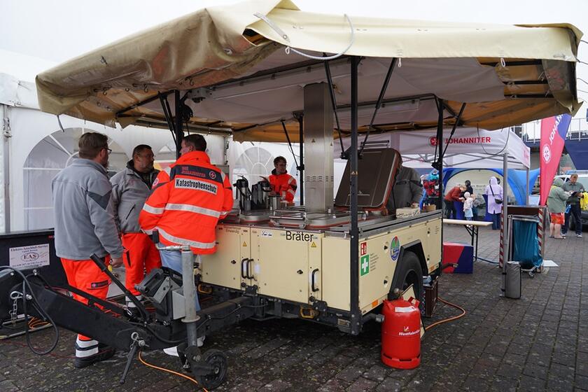 Man sieht eine Art mobile Küche, davor stehen drei Personen