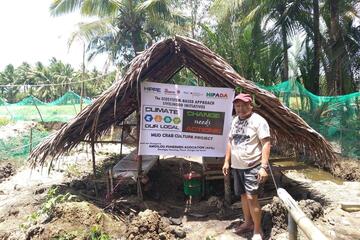 Ein Mann steht vor einer Holzhütte mit einem Banner des Projekts