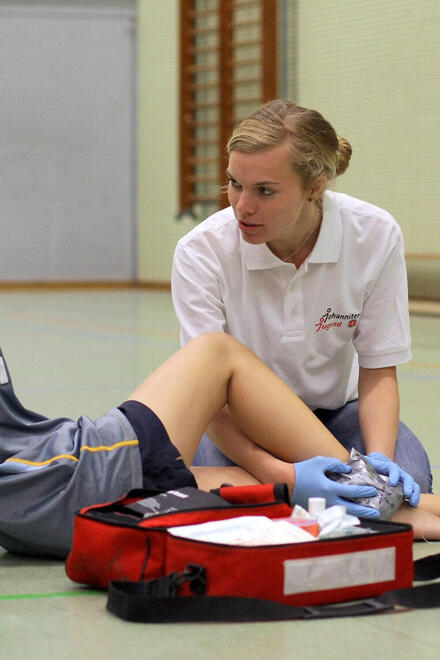 Ein Kind in Sportbekleidung sitzt in einer Sprothalle auf dem Boden. Eine junge Frau in Johanniter-Belkleidung versorgt sein Bein. 