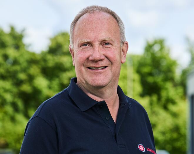 Ein Mann in mittleren Jahre mit Halbglatze und blauem Polo-Shirt. Im Hintergrund sind Bäume