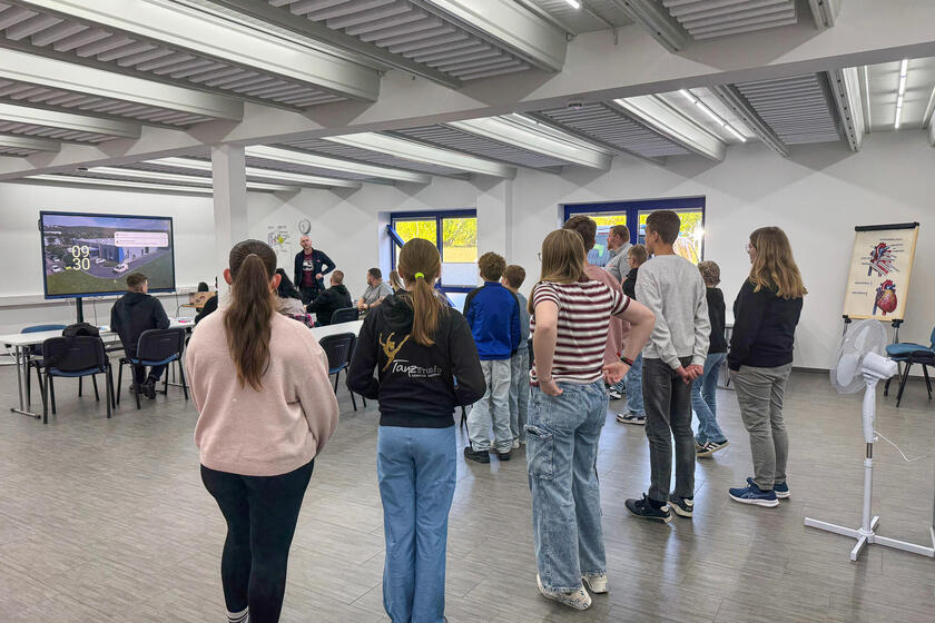 Jungen und Mädchen stehen in einem Lehrsaal. Hinter ihnen sitzen Personen an Tischen und ein Mann steht und spricht zu ihnen
