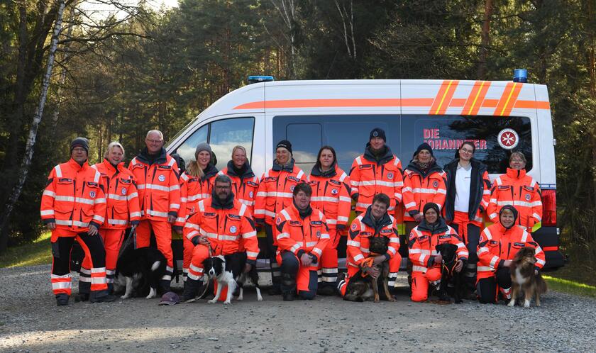 Gruppe von etwa 16 Personen in leuchtend orangefarbener Einsatzkleidung der Johanniter steht mit mehreren Hunden vor einem Einsatzfahrzeug im Wald. Das Fahrzeug trägt das Johanniter-Logo und ist mit blauen Rundumleuchten ausgestattet. 