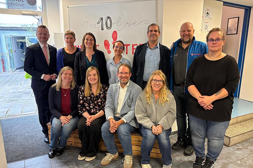 Mitarbeitende und Gäste der Johanniter feiern 10 Jahre Integrationszentrum „First Steps“ bei einem Gruppenfoto im Eingangsbereich, im Hintergrund ein Whiteboard mit der Aufschrift „10 Jahre First Steps“.