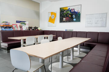 Aufenthaltsraum mit langen Holztischen und umlaufender brauner Polsterbank.