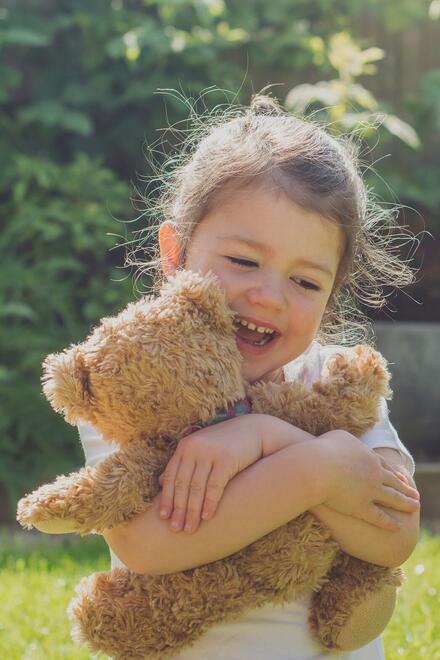Ein lachendes Kind sitzt im Garten und umarmt fröhlich einen braunen Teddybären im Sonnenschein.