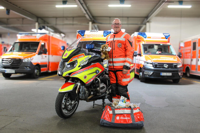 Ein Mann mit Johanniter-Motorradkleidung neben einem Motorrad. Er hält einen Teddy in der Hand. Vor ihm steht eine Tasche mit Teddys im Hintergrund Rettungswagen.