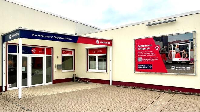 Ein gelbes Flachdachgebäude mit einer doppelseitiges gläsernen Eingangstür die überdacht ist an der rechten Hauswand ein Plakat mit Johannitern drauf.